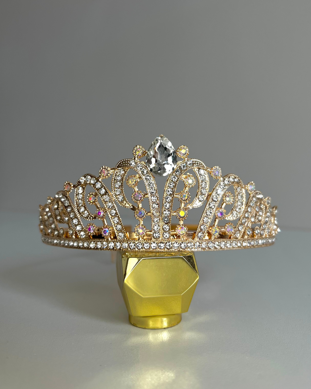 A golden tiara crown with silver rhinestones displayed on a yellow stand against a grey background.