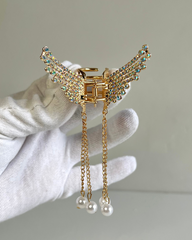 A golden hair claw clip designed to resemble angel wings with shimmering crystals and gold-toned chains dangling lustrous faux pearl accents.