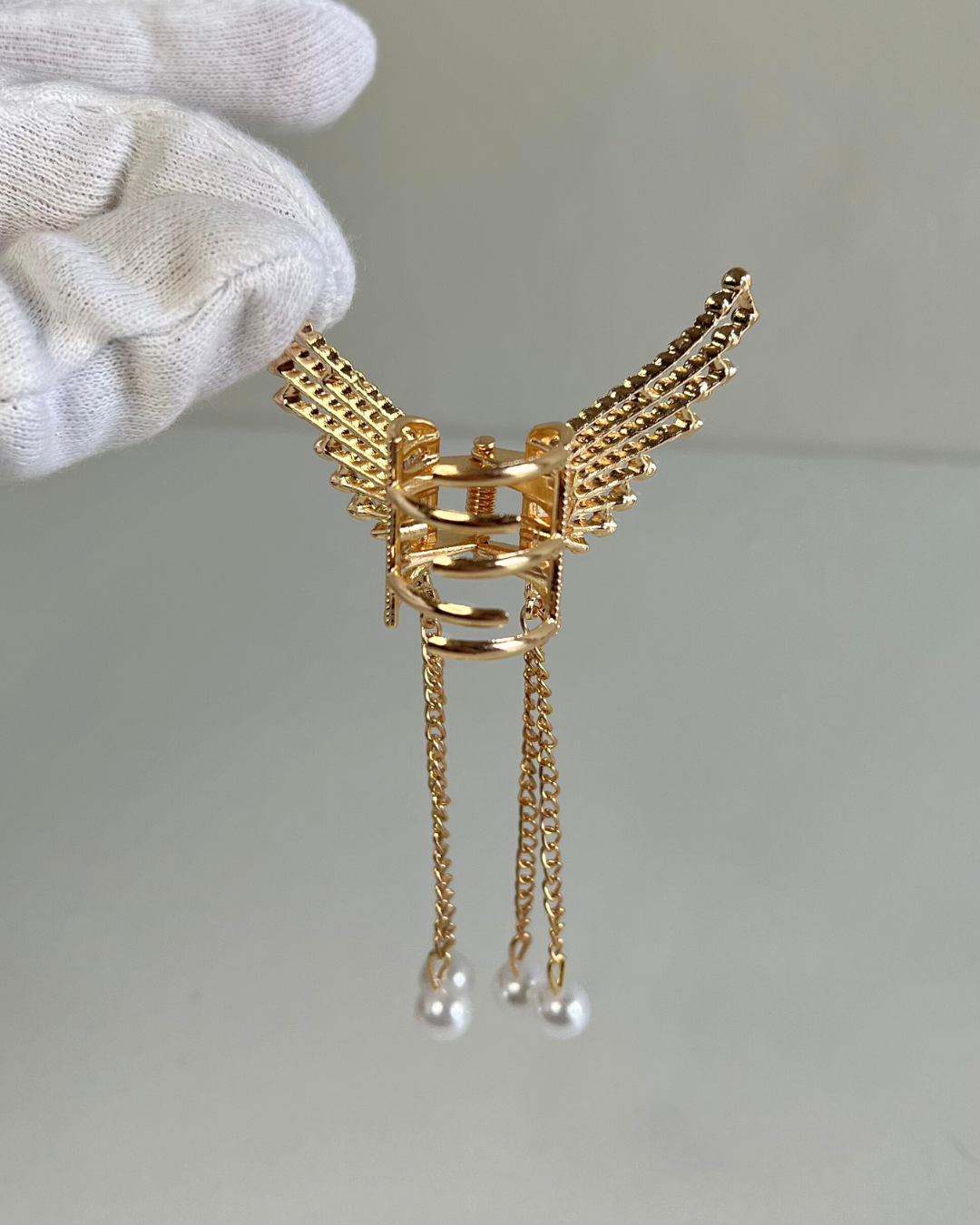 Gold winged earring with pearl drops held by a gloved hand on a neutral background.