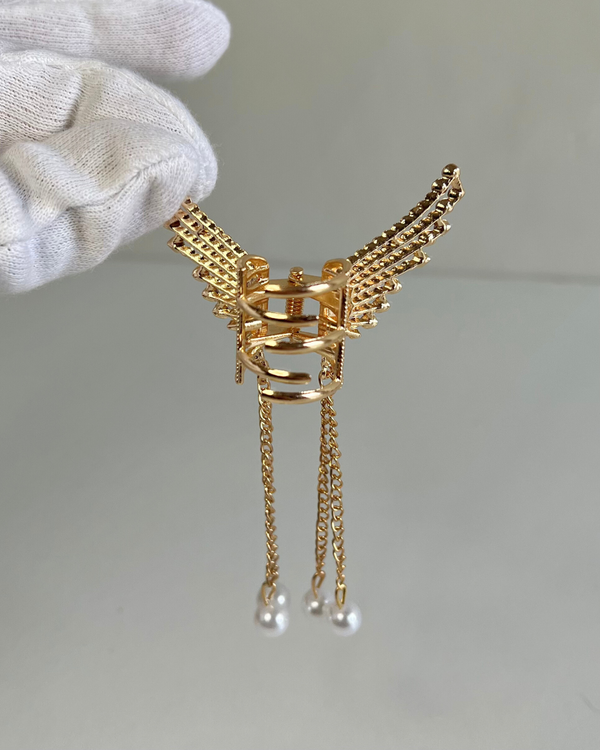 Gold winged earring with pearl drops held by a gloved hand on a neutral background.