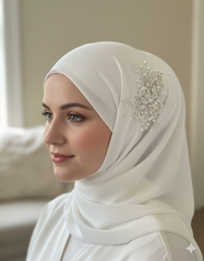 Woman wearing a white hijab with a decorative brooch, sitting indoors.