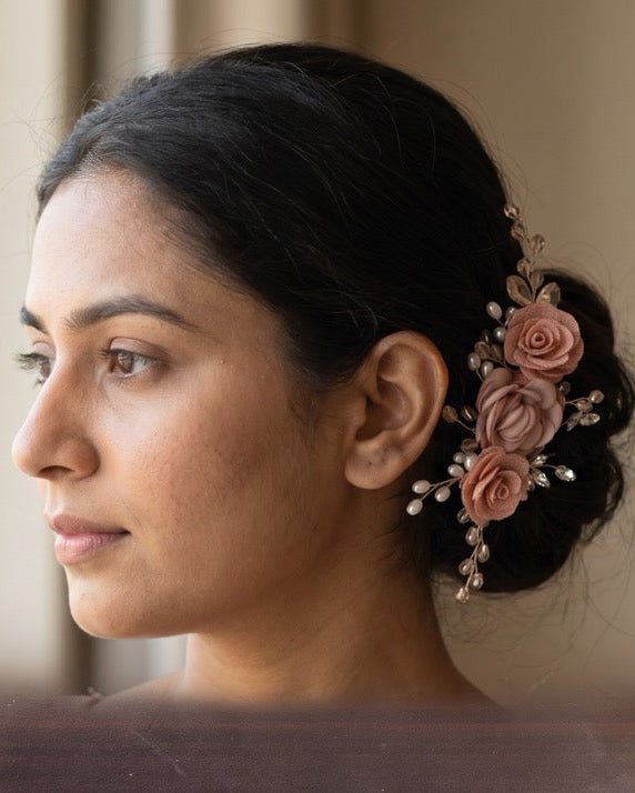 A woman in profile wearing a floral hair accessory with pink roses and small pearl-like beads in her dark hair, set against a softly blurred indoor background.